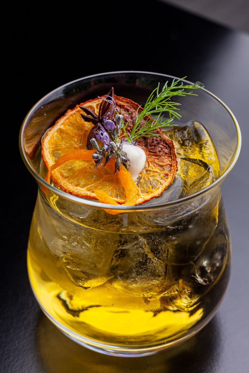 Cocktail giallo con arancia caramellata, rosmarino e petali viola