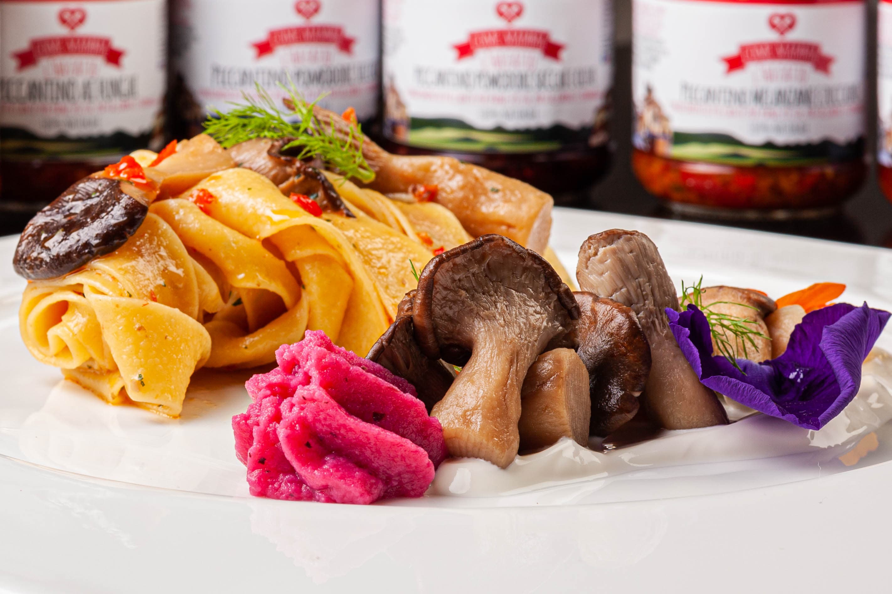 Pappardelle ricche, vasetti del brand sullo sfondo, mousse di barbabietola e fiori magenta — il condimento incontra la cucina